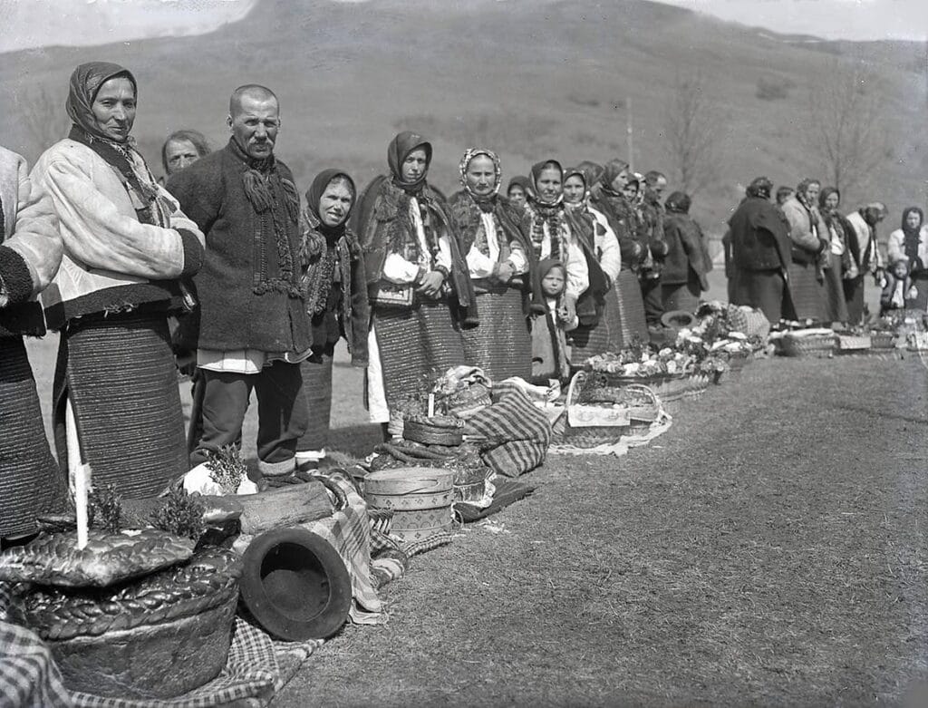 Українські селяни в автентичному одязі стоять у черзі біля пасхальних кошиків на освячення біля церкви 100 років тому. Старовинне чорно-біле фото.